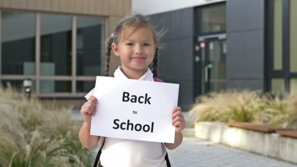 Nettes lächelndes kleines Schulmädchen in Uniform steht mit einem Plakat Back To School in der Nähe des Schulgebäudes. — Stockvideo