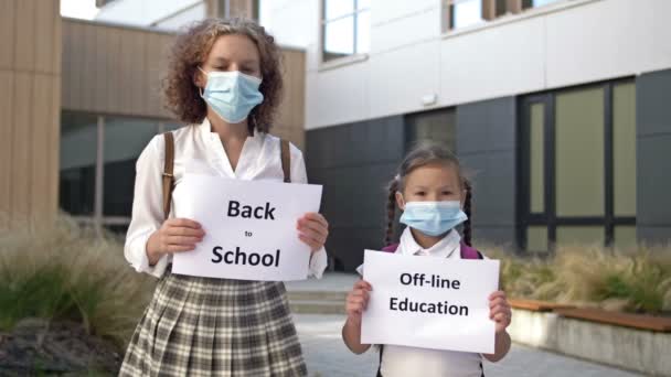 Två skolflickor som bär medicinska masker med plakat står framför skolbyggnaden. Affischen i händerna på en liten flicka OFFLINE UTBILDNING. Tonårstjej med affisch BAKA TILL SKOLA — Stockvideo