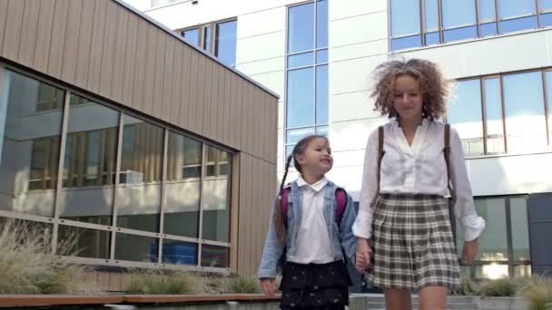 Dos colegialas de diferentes edades con mochilas van a la escuela, cogidas de la mano. La hermana mayor lleva a la más joven a la escuela. Regreso a la escuela. — Vídeo de stock