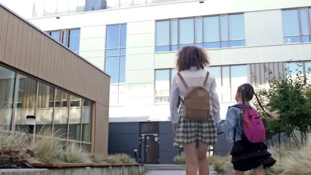 Sırt çantalı, farklı yaşlarda iki kız öğrenci el ele tutuşarak okula gidiyor. Büyük kız kardeş küçük olanı okula götürür. Arka plan. — Stok video