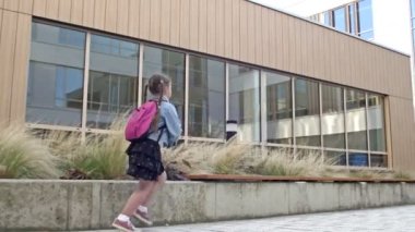 Bir ilkokul öğrencisi aceleyle okula gider. Okul binasının arka planında at kuyruklu ve sırt çantalı küçük bir kız koşuyor..