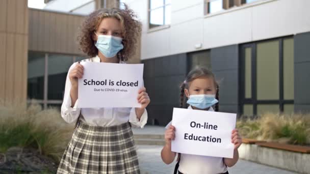 Två skolflickor som bär medicinska masker med plakat står framför skolbyggnaden. Affischen i händerna på en liten flicka ON-LINE UTBILDNING. Tonårstjej som håller i en affisch SKOLA ÄR CLOSED. — Stockvideo
