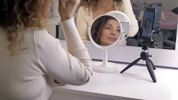 Un bloguero de belleza bien cuidado y de mediana edad habla sobre técnicas de maquillaje. Una mujer está sentada frente a un espejo y una cámara de video. — Vídeo de stock