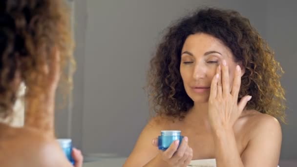 Hermosa mujer madura sosteniendo tarro de crema de piel para la cara. Mujer feliz aplicando crema hidratante anti-envejecimiento y mirando a la cámara. Belleza y tratamiento antienvejecimiento. — Vídeo de stock