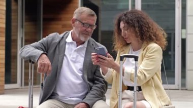 Bavulları olan güzel olgun bir çift bir bankta oturmuş uçuşlarını bekliyorlar. Bir adam ve bir kadın cep telefonunun ekranında ne gördüklerini tartışıyorlar..