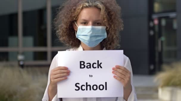 Tıp maskesi takan kız öğrenci okul binasının yakınında bir tabela tutuyor.. — Stok video