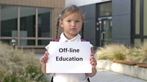 Серьезная маленькая школьница с косичками стоит с плакатом OFFLINE EDUCATION возле здания школы. — стоковое видео