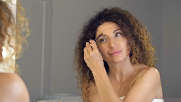 Gepflegte reife Frau kämmt ihre Augenbrauen mit Brauenbürste vor dem Spiegel. — Stockvideo