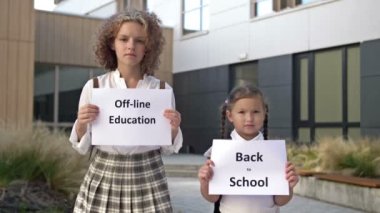 Okul binasının önünde pankartlı iki kız öğrenci duruyor. Okula geri dönen küçük bir kızın elinde bir poster. - Ne? Genç bir kızın elinde çevrimdışı eğitim afişi var..