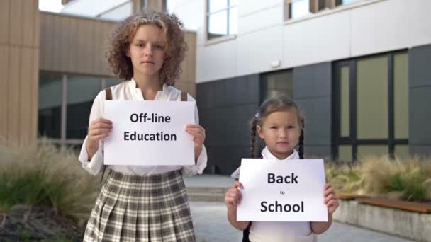 Två skolflickor med plakat står framför skolbyggnaden. En affisch i händerna på en liten flicka BAKA TILL SKOLA. En tonårig flicka innehar en affisch OFFLINE UTBILDNING. — Stockvideo