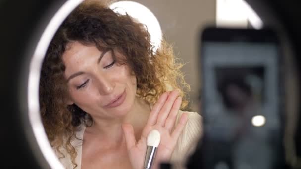 Beautiful, middle-aged beauty blogger talks about the types of makeup brushes and their purpose. Woman sitting in front of a mirror in which a video camera is reflected. — Stock Video