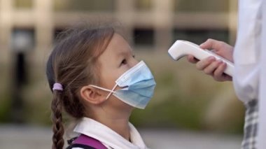 Koruyucu maskeli küçük bir kız öğrenci sıcaklığı kontrol ediyor. Salgın sırasında alınan önlemler.