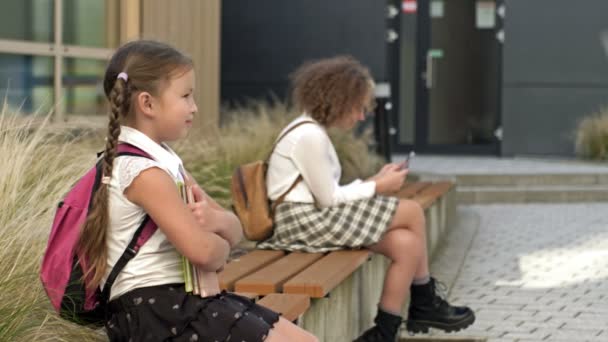 Dos colegialas de diferentes edades están sentadas en un banco en el patio de la escuela. La niña presiona los libros en su pecho, el anciano tiene un teléfono inteligente en sus manos. — Vídeos de Stock