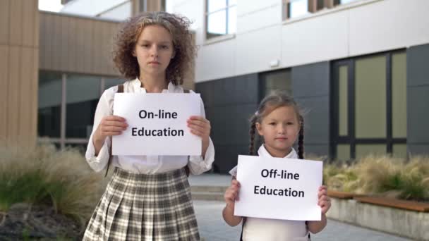 Dua anak sekolah dengan plakat berdiri di depan gedung sekolah. Sebuah poster di tangan seorang gadis kecil OFFLINE EDUCATION. Seorang gadis remaja memegang poster ONLINE EDUCATION. — Stok Video