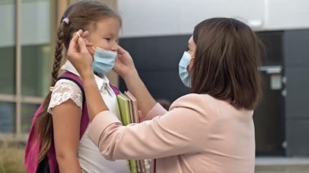 Eine junge Frau mit Schutzmaske setzt ihren Töchtern eine Maske auf, bevor sie die Schule betritt. Vorsichtsmaßnahmen während der COVID-19-Pandemie. — Stockvideo