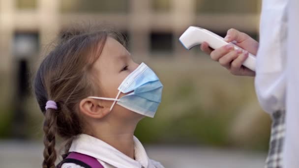 Klein schoolmeisje met een beschermend masker controleert de temperatuur. Voorzorgsmaatregelen tijdens een pandemie. — Stockvideo