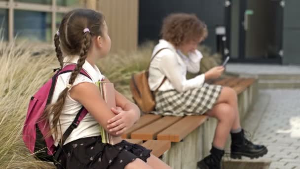 Två skolflickor i olika åldrar sitter på en bänk på skolgården. Den lilla flickan trycker böckerna mot bröstet, den äldre har en smartphone i sina händer. — Stockvideo