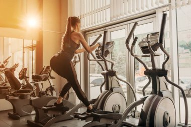Genç, formda bir kadın, spor salonunda eliptik bir eğitmen kullanıyor.