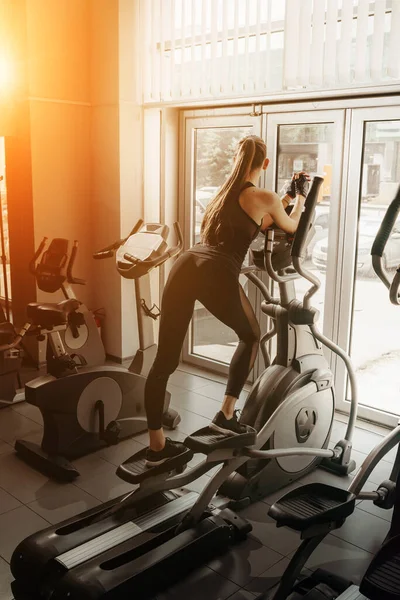 Genç, formda bir kadın, spor salonunda eliptik bir eğitmen kullanıyor.