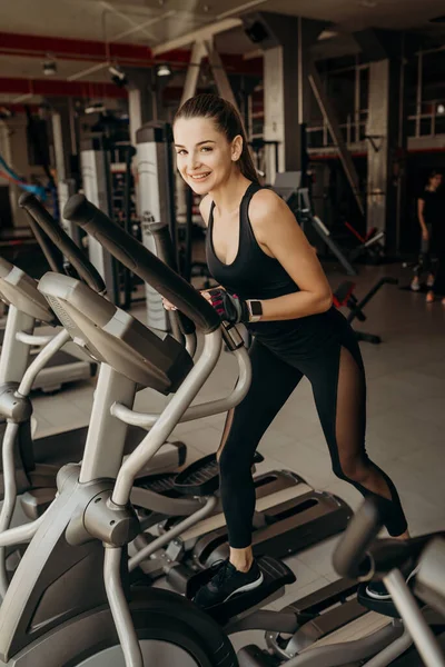 Genç, formda bir kadın, spor salonunda eliptik bir eğitmen kullanıyor.