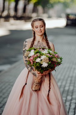 İki örgülü, güzel, uzun saçlı bir gelinin portresi. Elinde koca bir buket düğün çiçeği olan bir kız. Dövmeli kadın çok güzel pembe bir gelinlik giymiş..