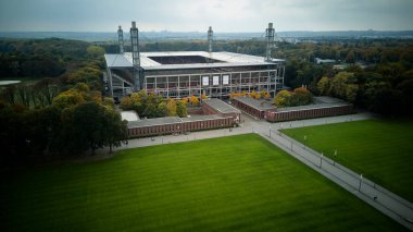 Cologne, Germany - 12 October, 2025: Wide lawn in front of a modern football stadium in Muengersdorf. High drone panorama of a square football stadium framed by trees, walkways and training fields in an urban park. Clear geometry, daylight