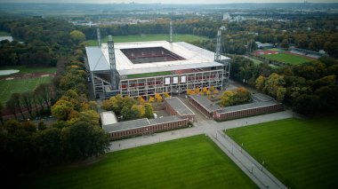 Cologne, Germany - 12 October, 2025: Symmetrical aerial view of a modern Muengersdorf football stadium aligned with a central walkway and lawns, set within a forested urban park. No people.