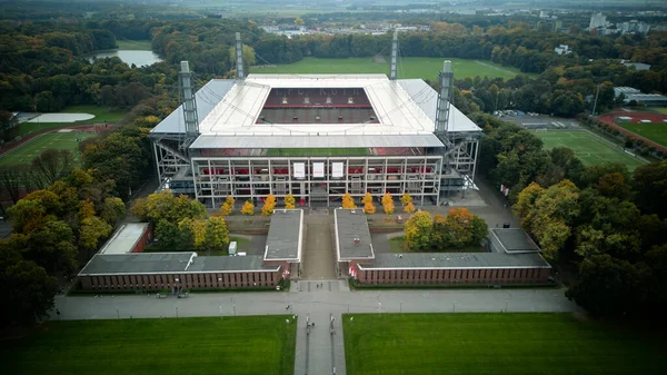 Cologne, Germany - 12 October, 2025: Symmetrical aerial view of a modern Muengersdorf football stadium aligned with a central walkway and lawns, set within a forested urban park. No people.