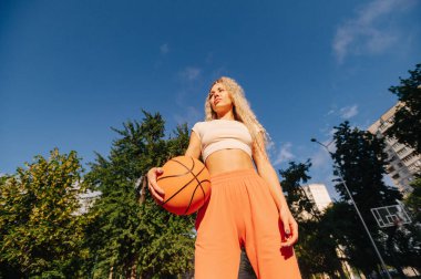 Kendine güvenen sporcu bir kadın, açık mavi gökyüzünün altında basketbol topunu tutuyor. Düşük açılı çekim gücü ve motivasyonu gösteriyor..