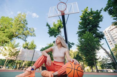 Sepetin altında oturan kadın oyuncu, el üstünde, yana bakıyor, canlı yeşil çevre ve açık gökyüzü, yaşam tarzı spor portresi.