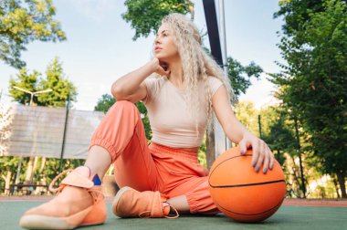 Kapalı alanda basketbol oynayan bir kadın manzarası, ılık yaz ışığında rahat bir an, modern sokak sporu sahnesi..