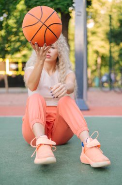 Sahada otururken basketbol oynayan genç bir kadın, hareketli bir an, parkta sokak sporu yaşam tarzı..