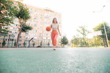 Basketbolcu kız boş şehir sahasında kameraya doğru yürüyor. Geniş ön planda ve parlak sabah ışığında minimum bileşim.