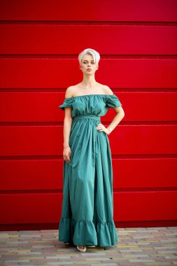 Kırmızı duvarın karşısında duran, omuzları açık, deniz mavisi bir elbise giymiş zarif genç bir kadın. Güçlü renk kontrastlı ve temiz, modern arka planlı tam boy moda portresi.