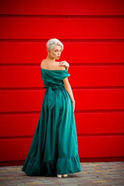 Kırmızı bir duvara karşı kameraya poz veren, omuzları açık, deniz mavisi bir elbise giymiş zarif bir kadın. Kalın renk kontrastlı şık sokak modası.