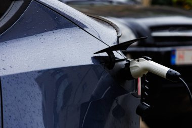 Yağmurlu havada dikiz aynasından elektrikli araba şarj oluyor. Yağmur damlaları, temiz enerji ve sürdürülebilir taşımacılığın sembolü olan şarj cihazına takılı elektrikli bir arabanın arka yakın çekimi.
