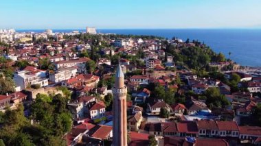 Aerial drone orbit around the historic Yivli Minaret in Antalyas Old Town, showcasing red-roofed houses, ancient streets and the Mediterranean coastline in vibrant daylight.