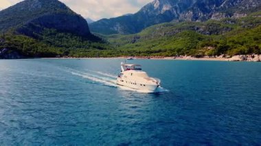 Aerial drone flying ahead of a luxury yacht cruising along the Antalya coast. Turquoise Mediterranean Sea with green mountains and rocky shoreline in the background.
