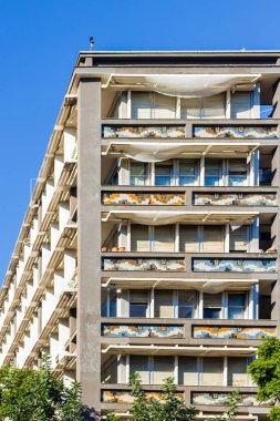 Çağdaş bir apartman bloğu desenli balkonları, geniş pencereleri ve havadar güneş gözlükleriyle. Yukarıdaki parlak mavi gökyüzü şehir yaşamını, sakin bir atmosferi ve cephe boyunca mimari detayları ön plana çıkarıyor..