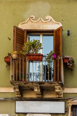 Yıpranmış yeşil cephedeki tuhaf bir balkonda ahşap panjurlar, çeşitli saksı bitkileri ve dekoratif korkuluklar bulunur. Penceredeki parlak yansımalar sıcaklık ve biraz Akdeniz cazibesi ekler..