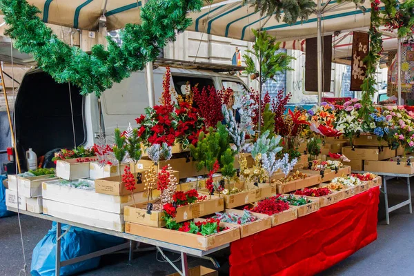 Çiçek aranjmanları ve dekoratif çelenklerle dolu hareketli bir açık hava tezgahı. Parlak kırmızılar, yeşiller ve beyazlar beyaz bir minibüsün yanında tatil vitrini oluşturuyor. Müşterileri gezmeye ve çiçek almaya davet ediyor..