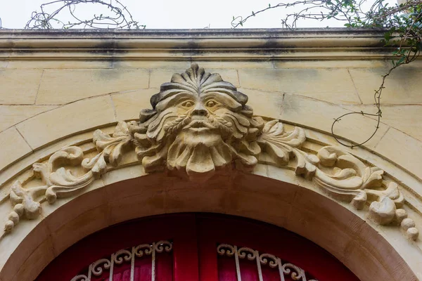 Kırmızı kapının üzerinde oyulmuş yüz heykeli ve asma detayları olan süslü taş kemer yolu.