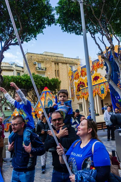 Cospicua, Malta - November 30th, 2025, Vibrant Street Parade With Blue And White Balloons, Flags, And Crowds Celebrating Festival