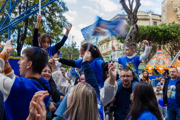 Cospicua, Malta - November 30th, 2025, Vibrant Street Parade With Blue And White Balloons, Flags, And Crowds Celebrating Festival