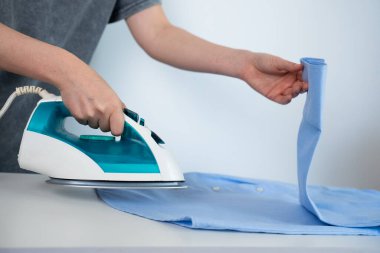 Person ironing a blue shirt with a modern iron on light background. Household and clothing care concept. 