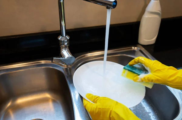 Hand in yellow glove rinses a white plate under running water in a stainless sink, everyday dishwashing, hygiene and kitchen routine.
