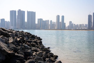  Modern skyscrapers of coastal city viewed from rocky beach under soft light. 