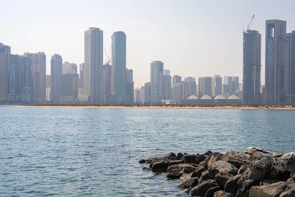 Modern skyscrapers mirrored in calm blue sea under hazy sky in coastal city. 