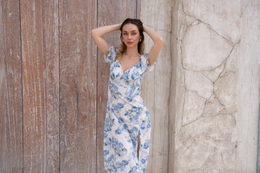 Açık mavi çiçekli güzel bir kadın ahşap kapının yanında poz veriyor, yumuşak güneş ışığı ve nötr renkler huzurlu ve zarif bir his yaratıyor.. 