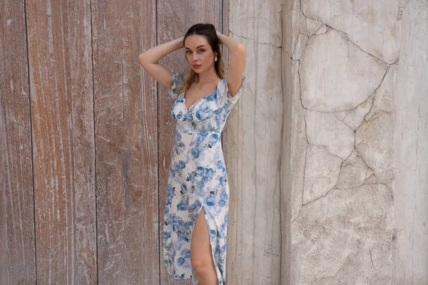 Saçlarını gün ışığı altında düzelten güzel bir kadın, desenli duvarın yanında çiçekli bir elbise giyiyor. Otantik ifadeli yumuşak bir yaz havası.